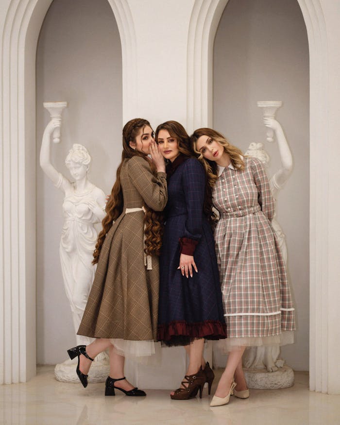 Three women in vintage dresses pose elegantly indoors with classical sculptures.